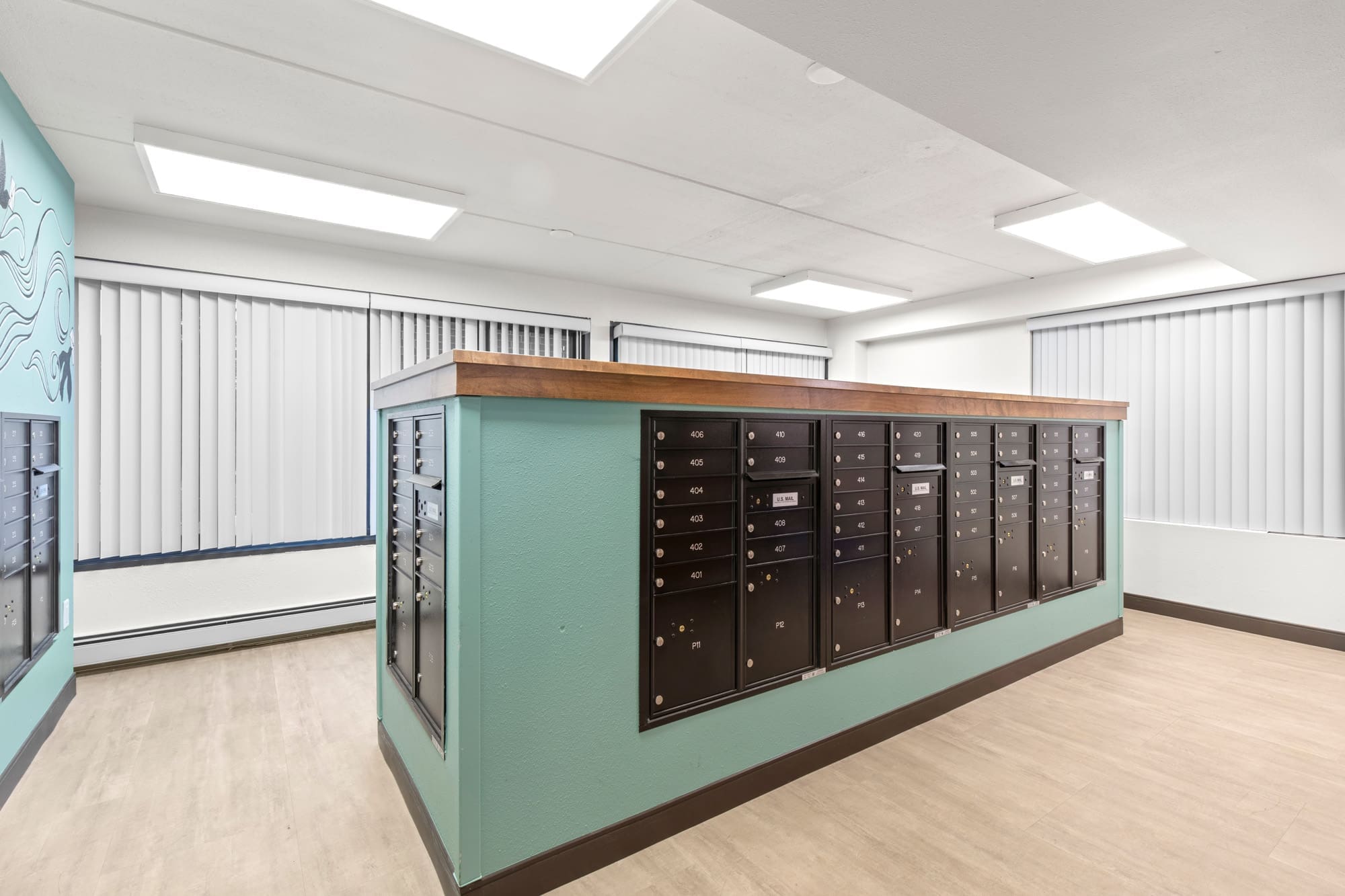 Modern commercial mailroom featuring teal walls, wood trim, black mailboxes, ample lighting, and windows with vertical blinds.
