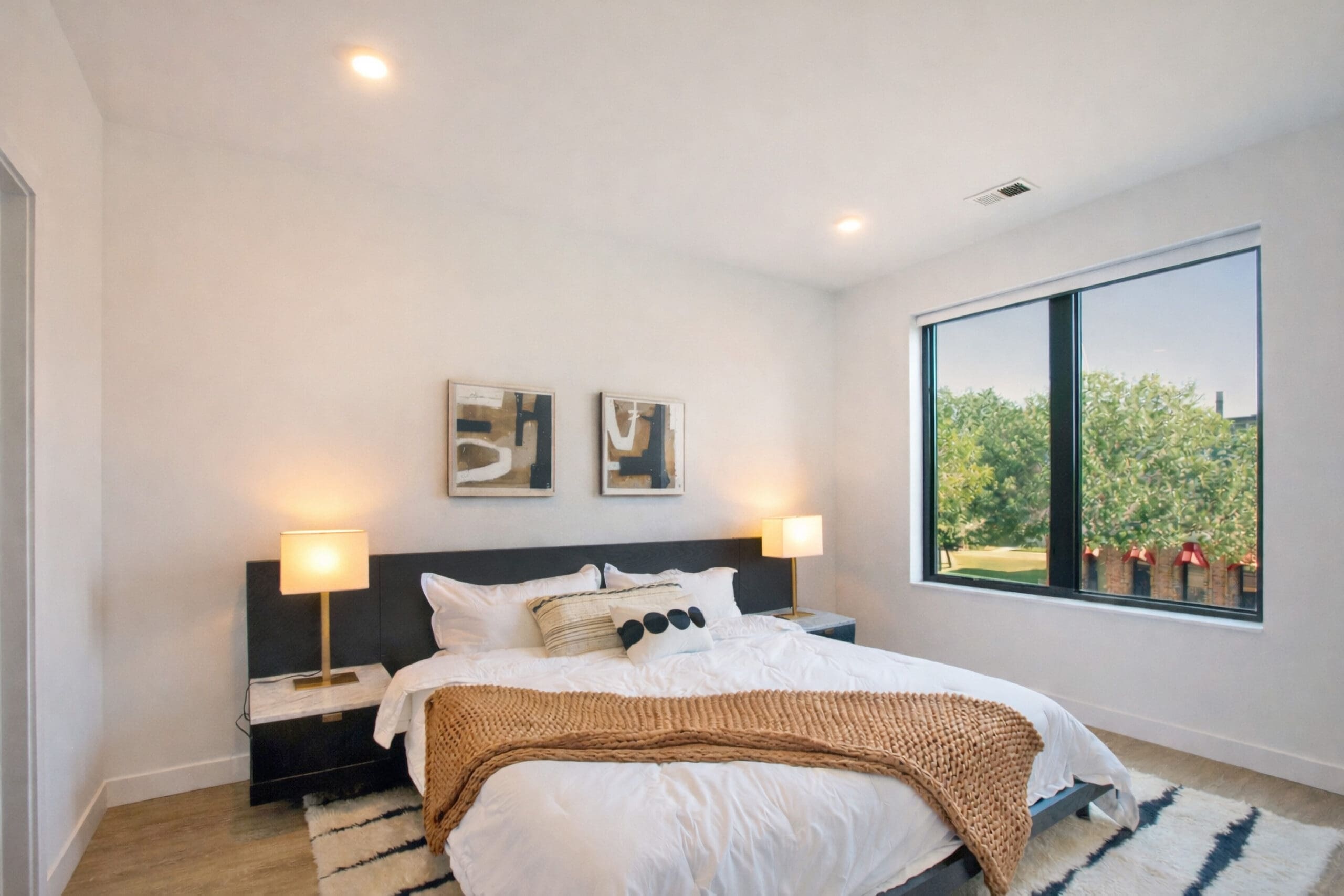 Contemporary residential bedroom featuring a spacious bed, bedside tables with lamps, abstract artwork, window view, and textured rug.