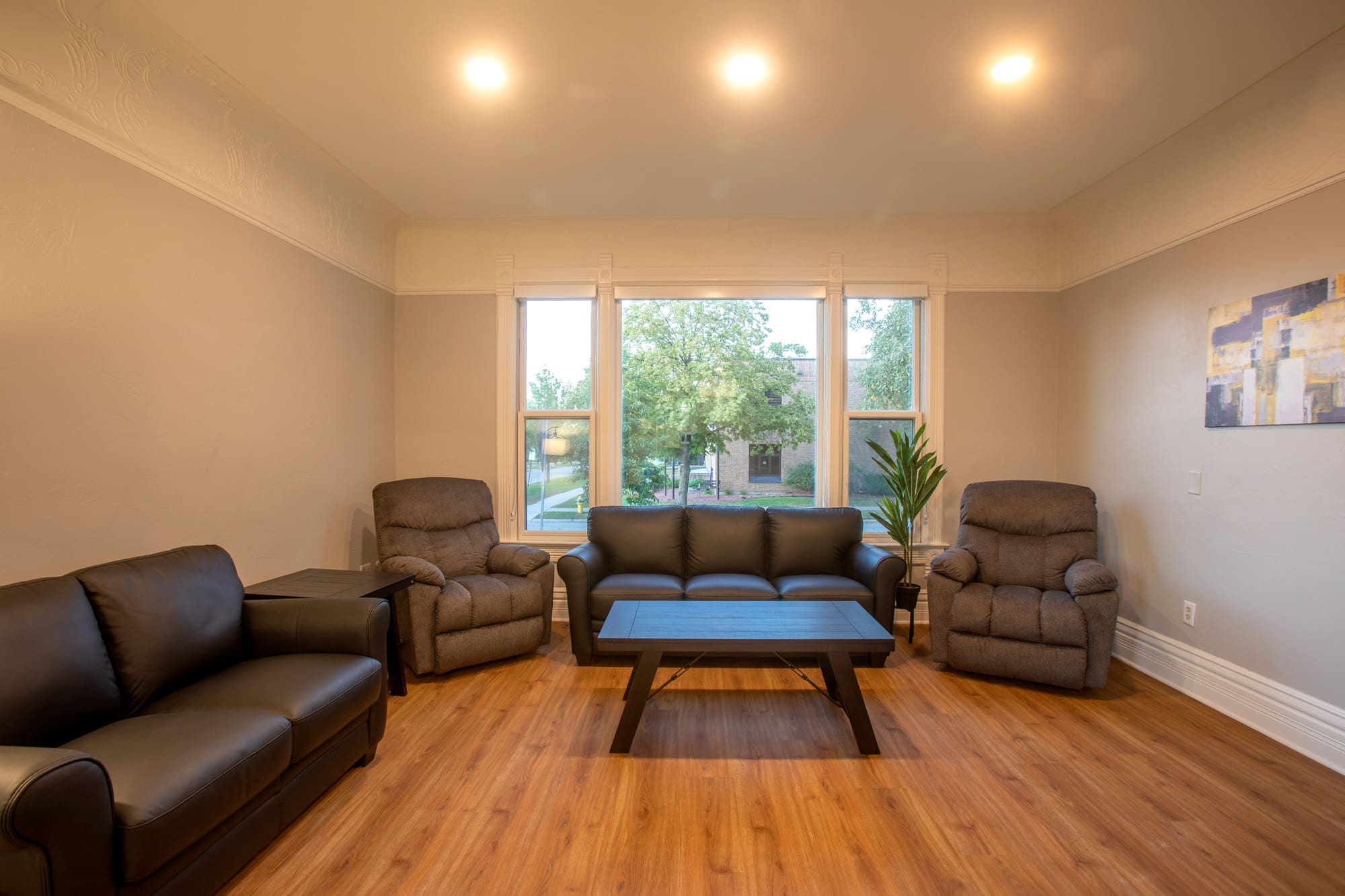 Bright residential living room featuring two recliners, a sofa, wood coffee table, potted plant, wall art, and large windows.