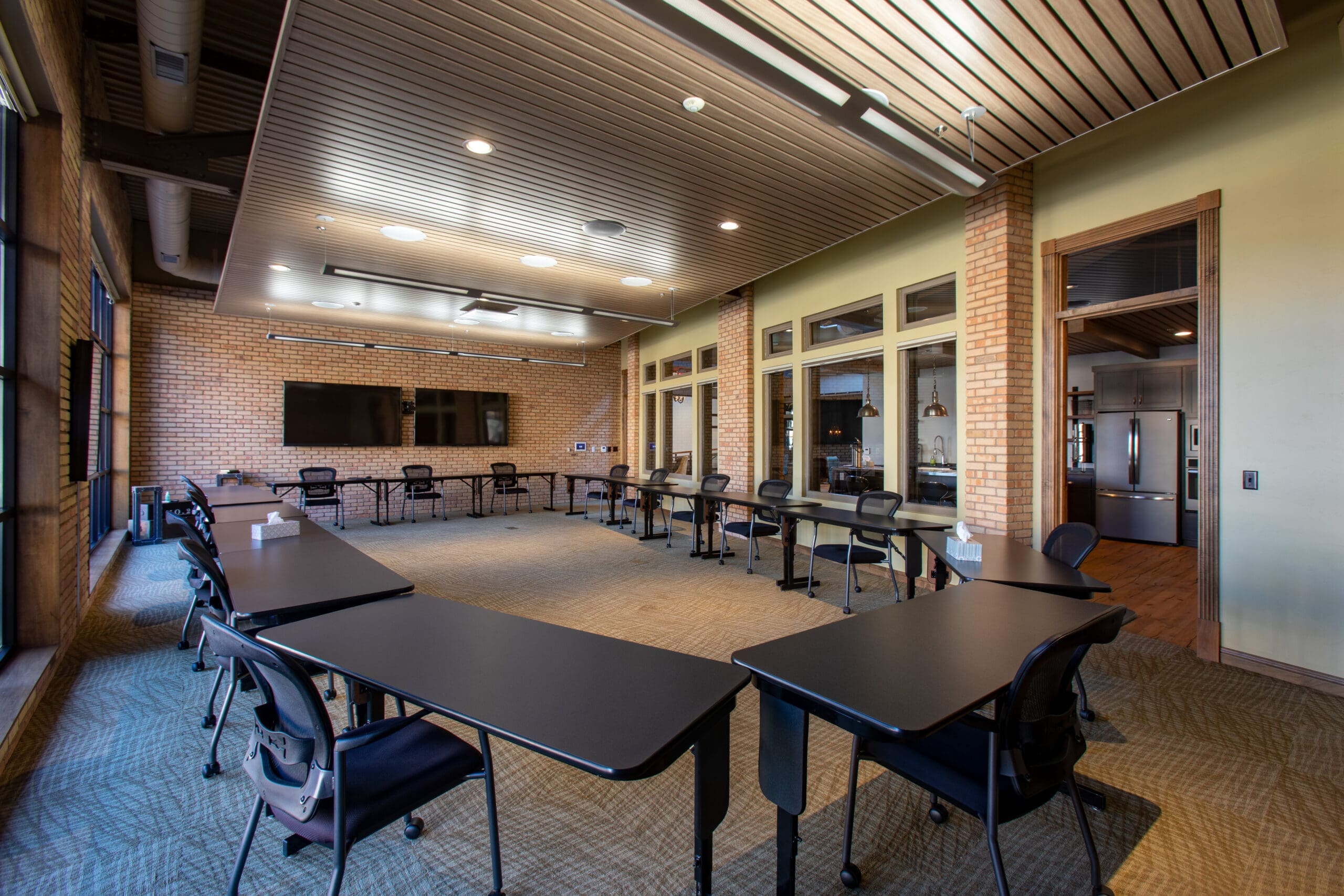 Commercial real estate conference room with U-shaped black tables, TV, brick accent wall, carpeted floor, and large sunlit windows.