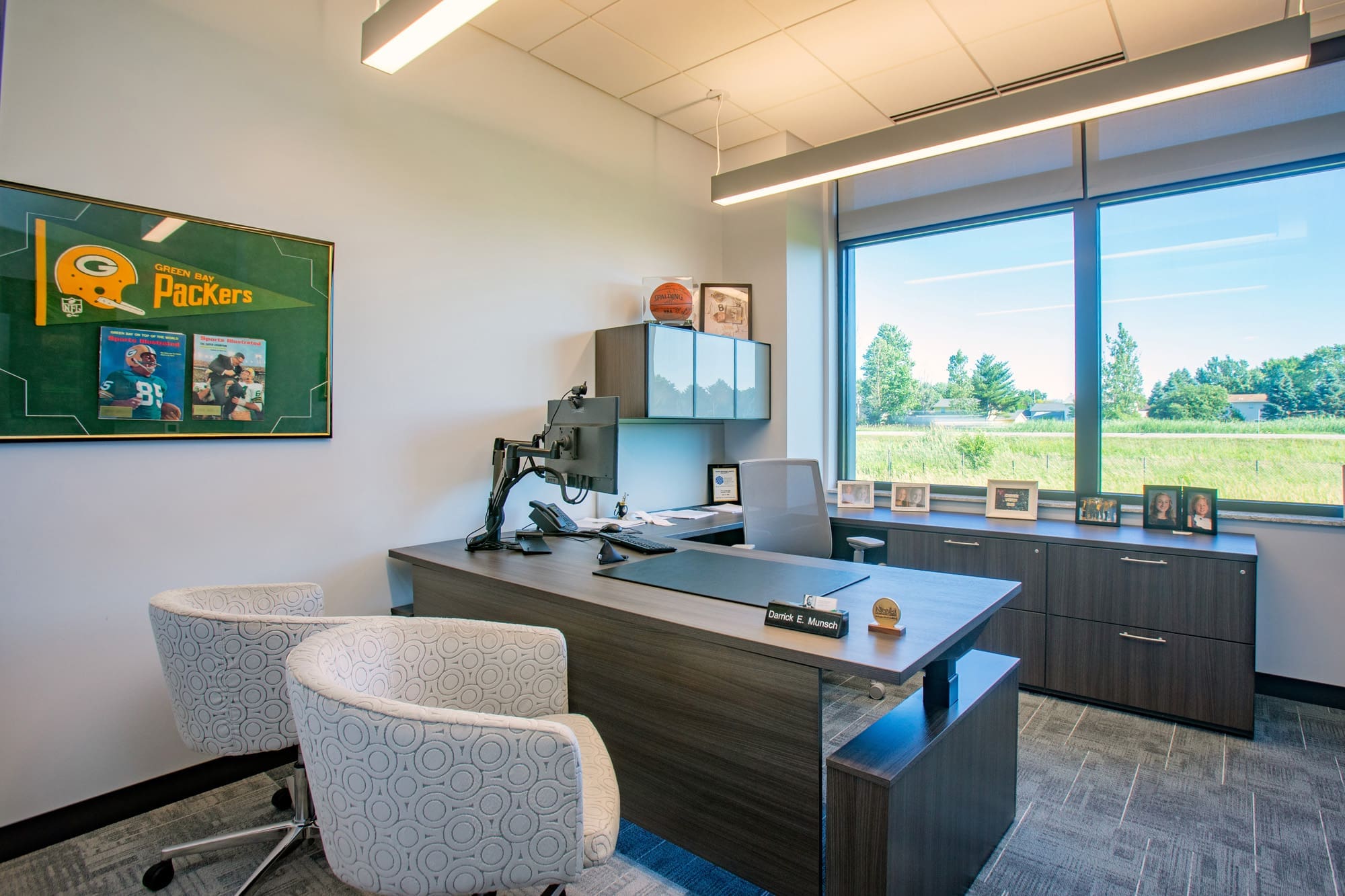 Spacious commercial office with large desk, two chairs, wall decor, cabinet photos, and big window overlooking landscaped grounds.