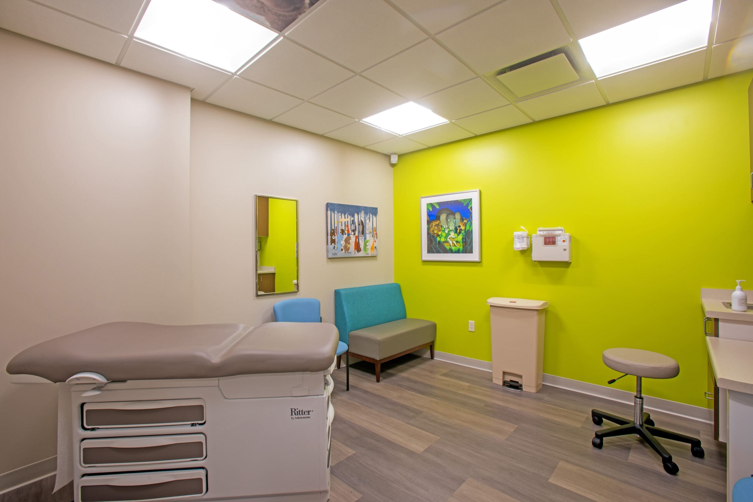 Commercial medical office exam room featuring an exam table, modern seating, equipment, bright walls, and decorative artwork.