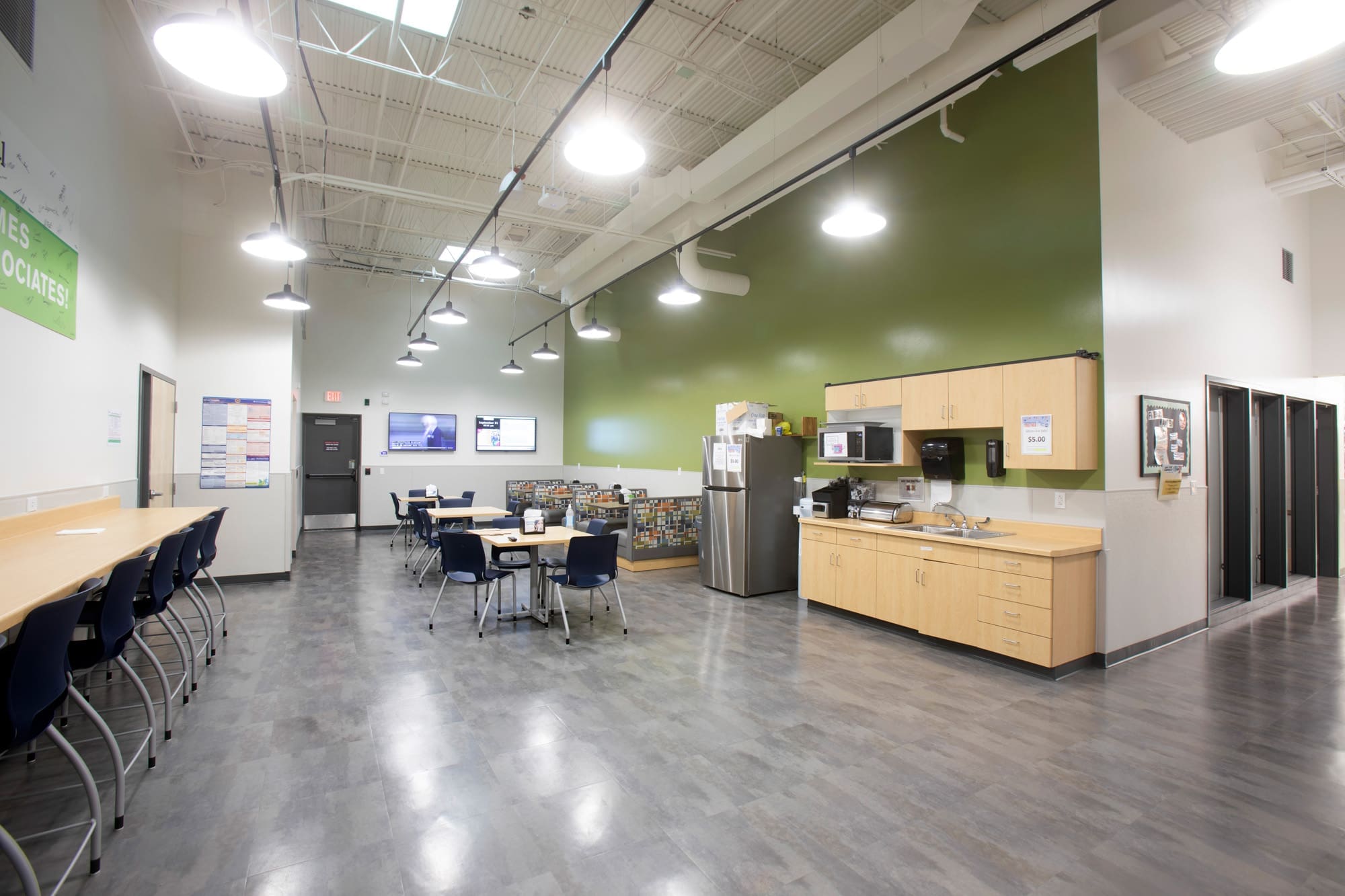 Spacious commercial break room with modern tables, chairs, kitchenette, microwave, coffee maker, and counter seating under bright lighting.