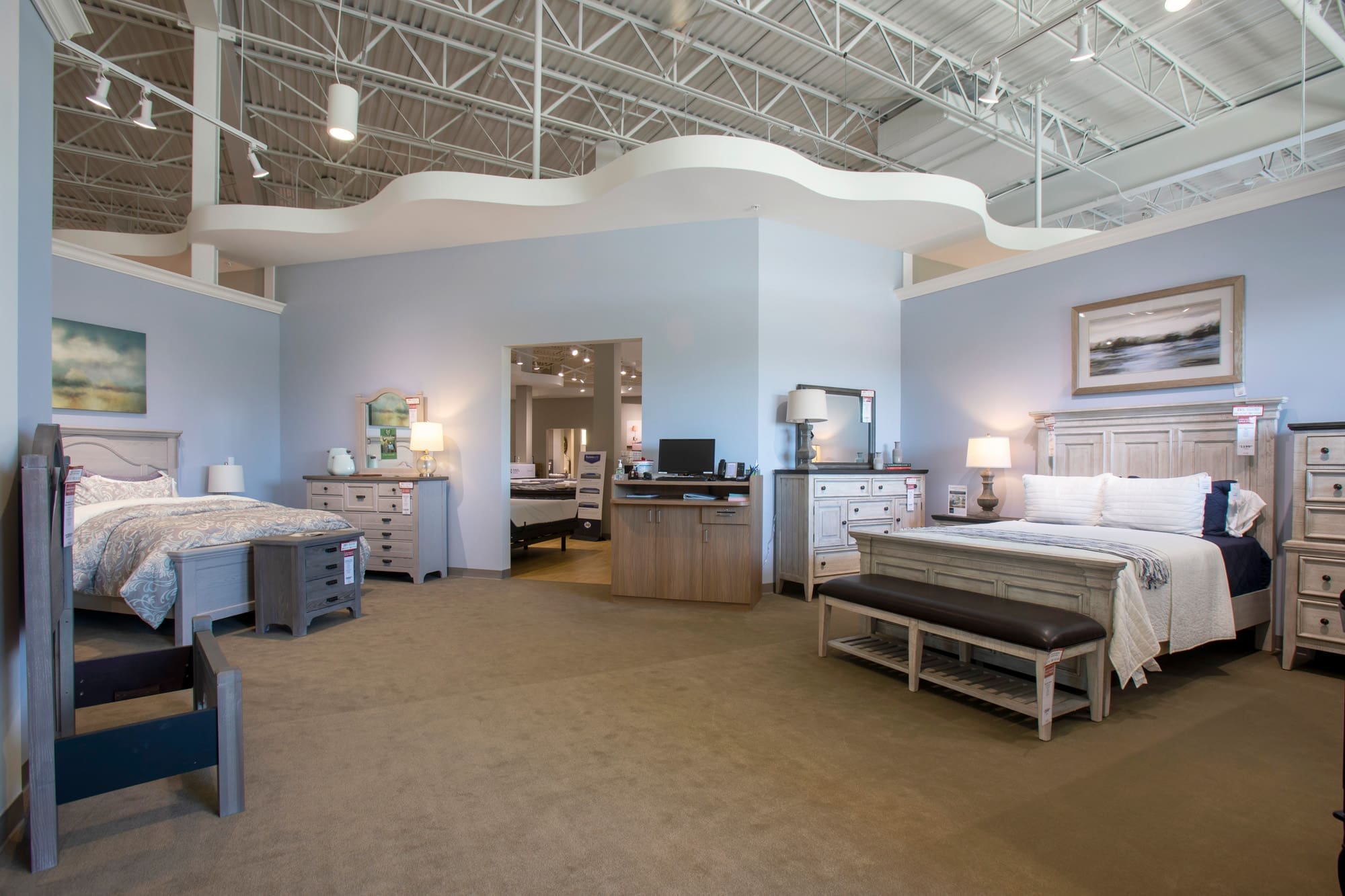 Residential showroom features two staged bedroom sets with beds, dressers, nightstands, and lofted ceiling for real estate display.