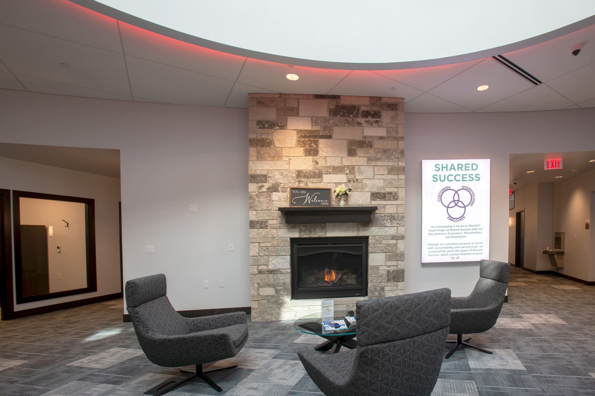 Commercial office lobby with modern gray chairs, round table, stone fireplace, and Shared Success wall display in bright setting.