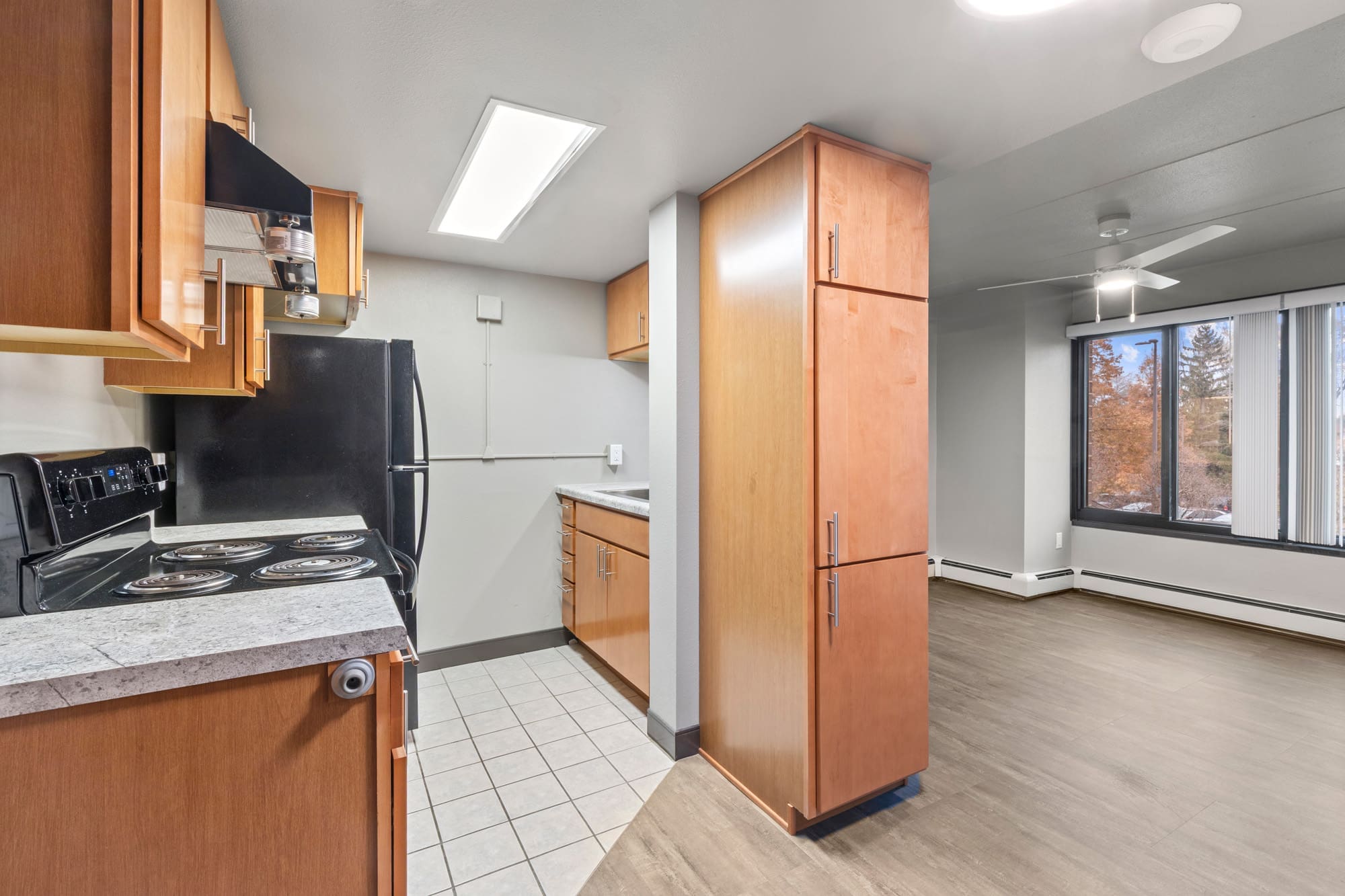 Residential kitchen with wood cabinetry, black appliances, tile flooring; open to living area with large window and wood-look floors.