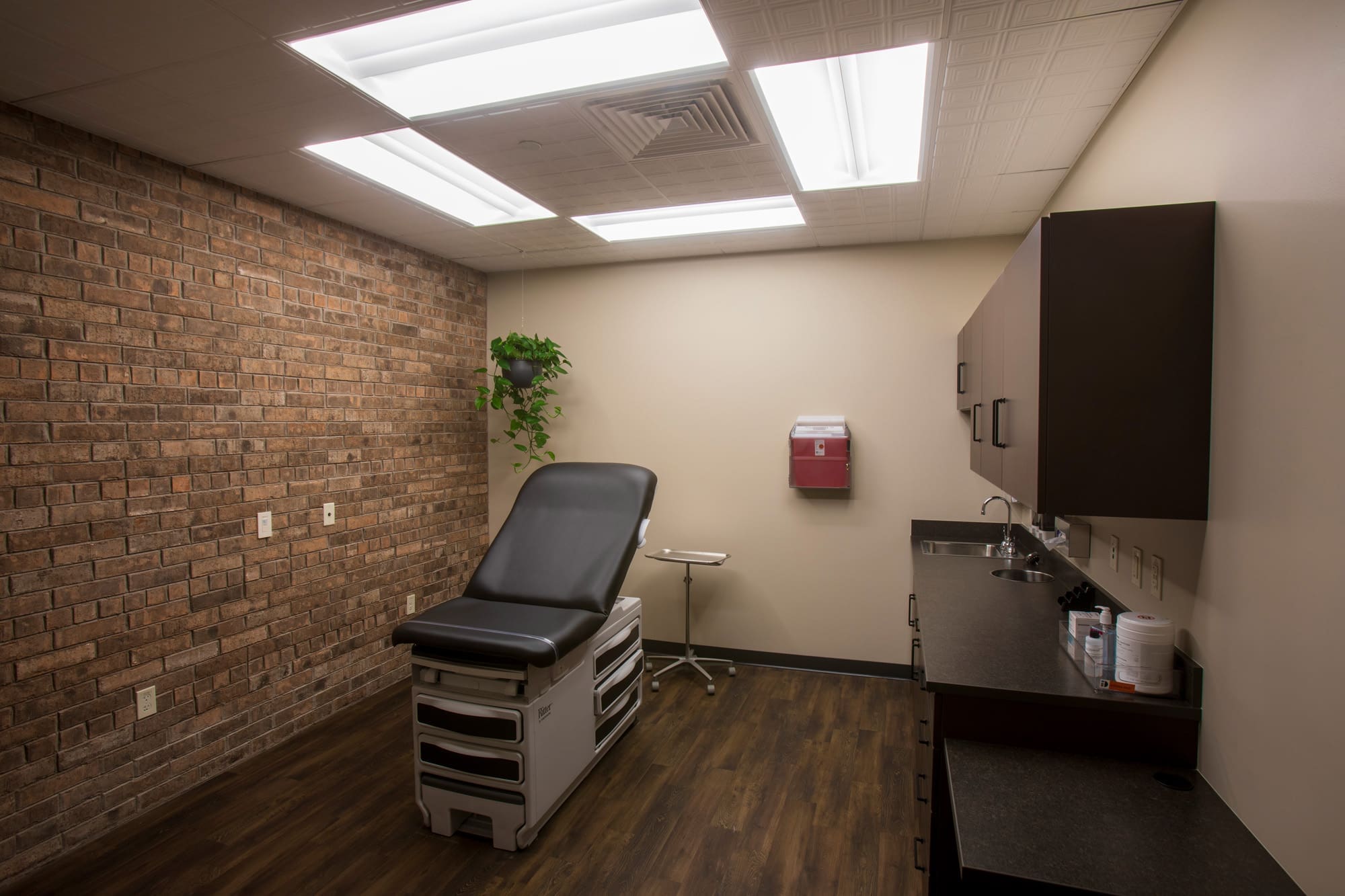 Commercial medical office examination room featuring brick accent wall, modern cabinetry, exam table, and natural light accents.