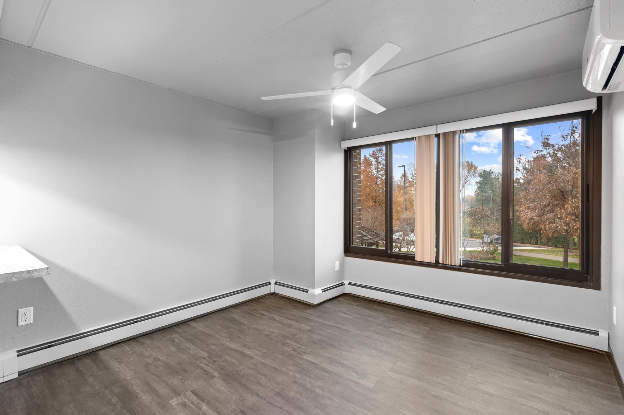 Spacious residential room with gray walls, wood flooring, ceiling fan, large window with blinds, and baseboard heating.