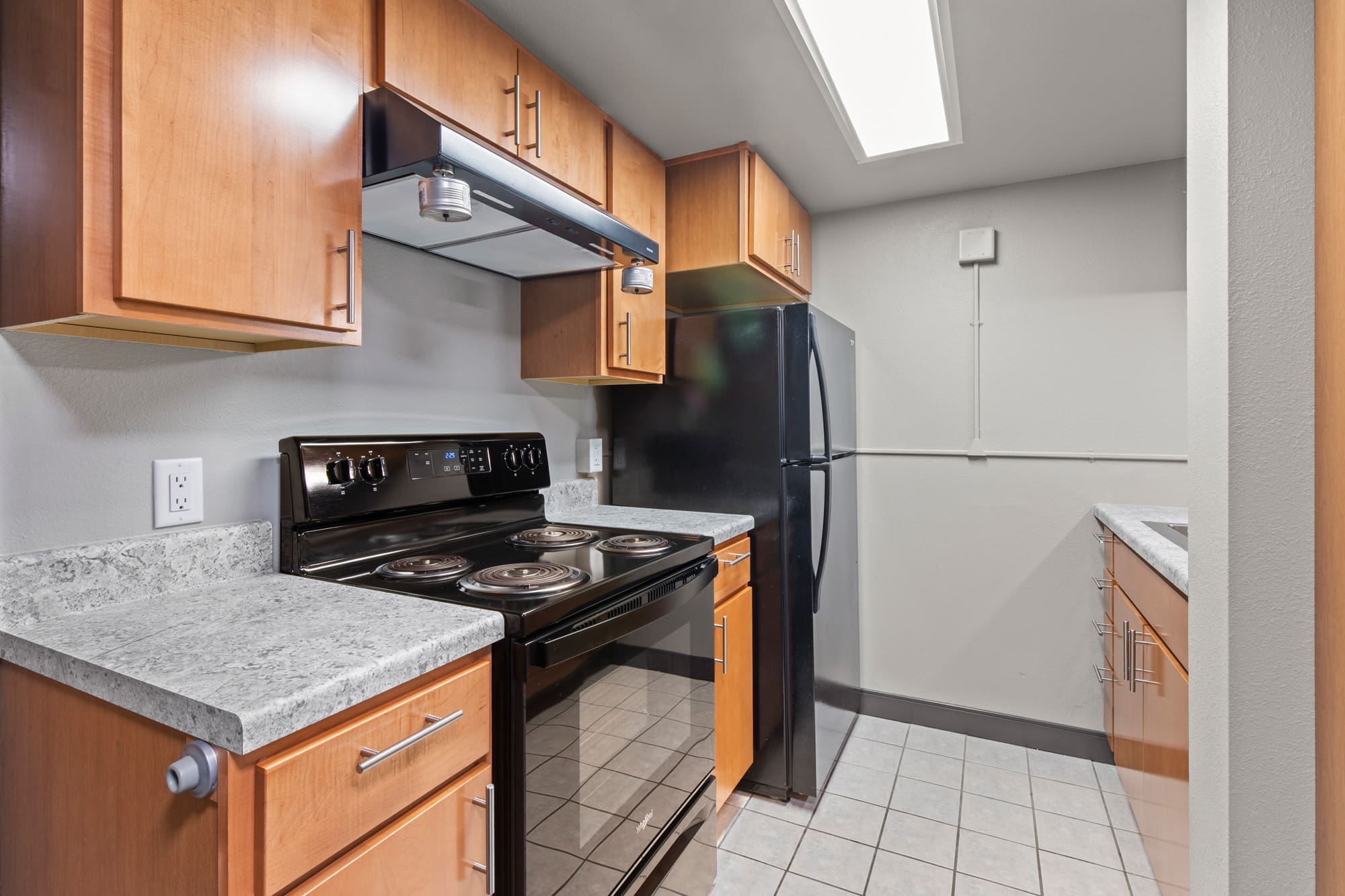 Residential kitchen featuring black appliances, wood cabinetry, marble-look countertops, and tiled flooring under bright ceiling lighting.