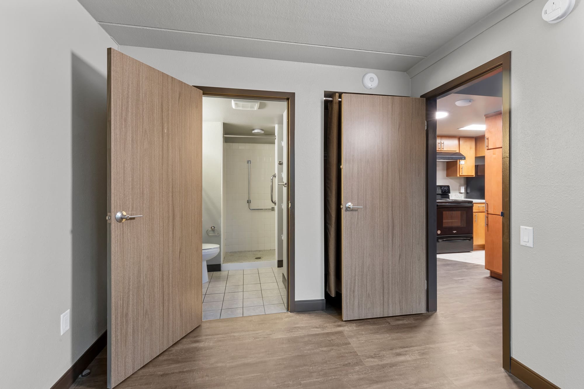 Residential property interior featuring a bedroom with ensuite walk-in shower, grab bars, closet space, and adjacent kitchen view.